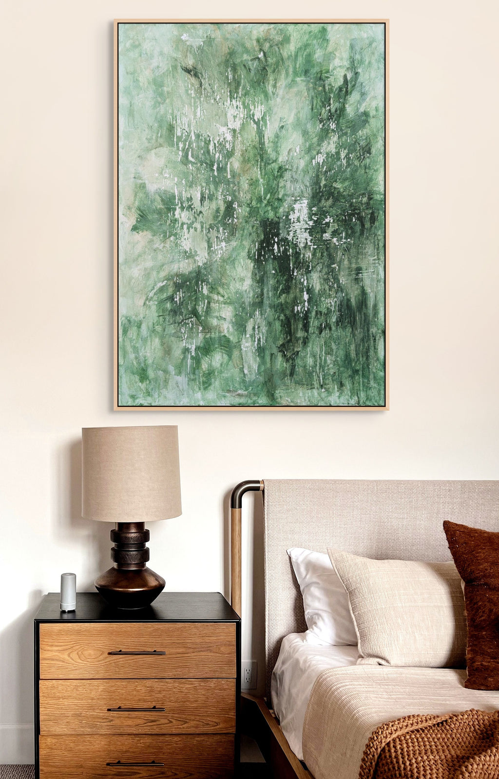 a bedroom with a large abstract painting hanging on the wall above a wooden nightstand with a lamp. The painting has a green and white color scheme, creating a serene and calming atmosphere in the room.