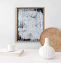 Framed abstract art piece on a wall with a white vase and woven basket below.