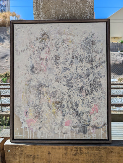 an abstract painting with a mix of white, gray, and pink colors, displayed in a wooden frame on a wooden surface.