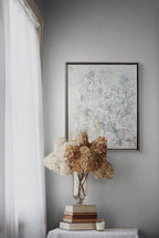 Decorative setup with a framed abstract painting, dried flowers, and books on a table against a gray wall.