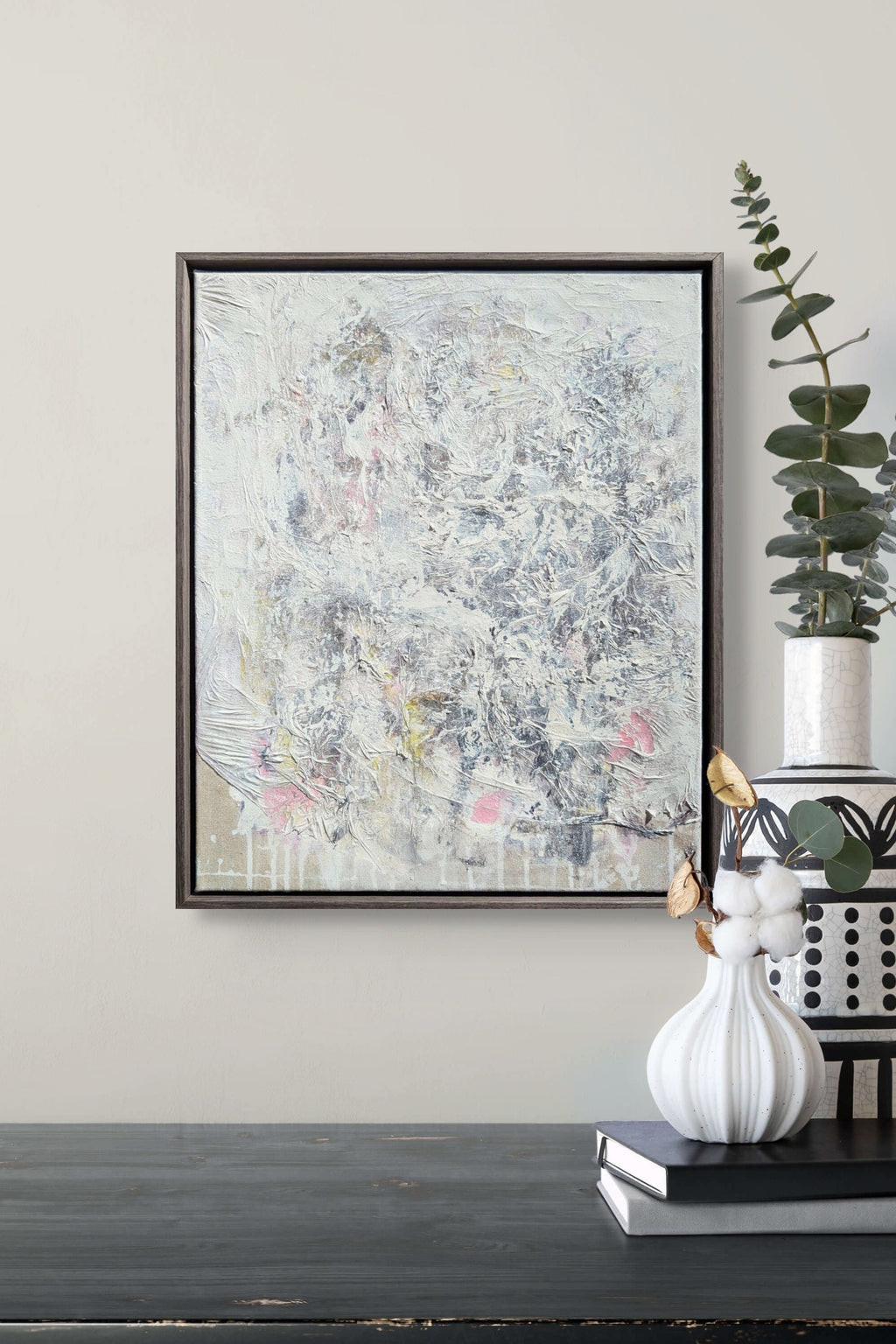 Abstract painting in a black frame on a wall above a dark surface with decorative items.