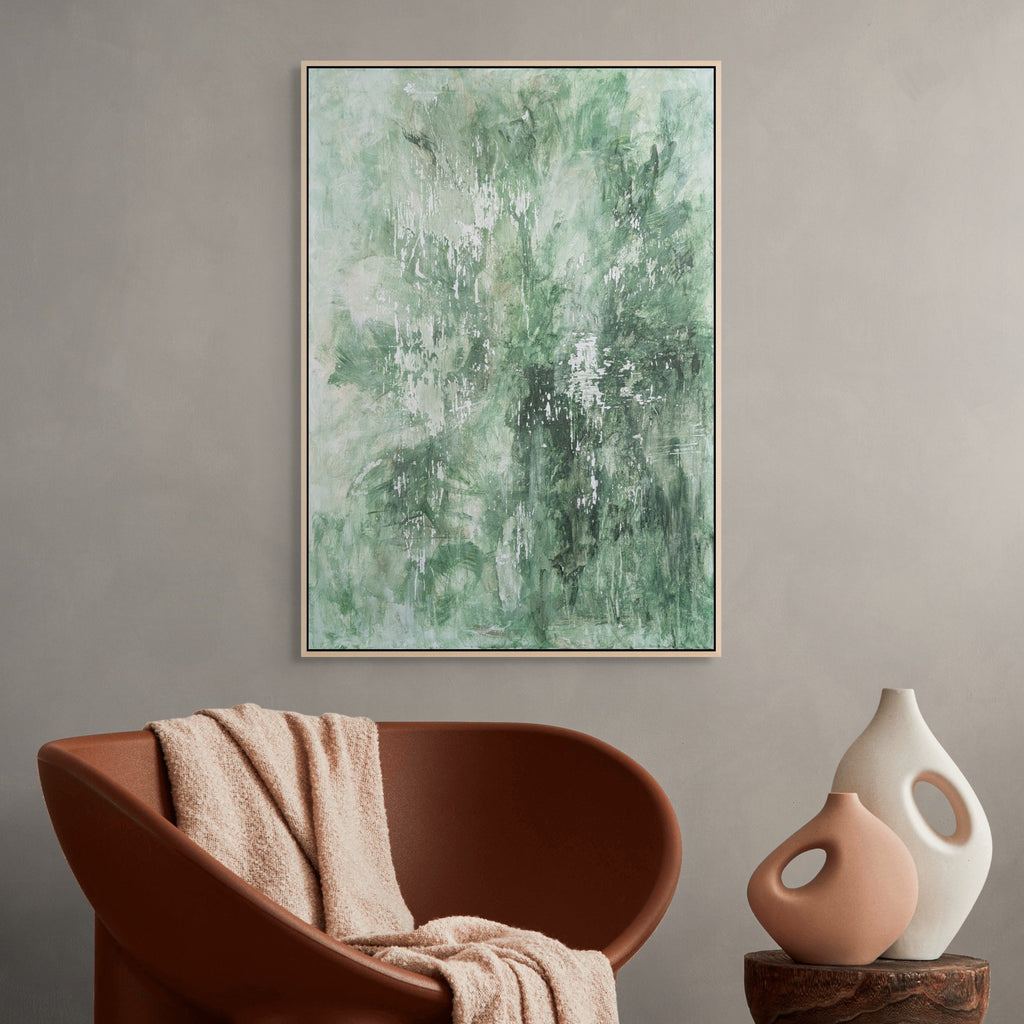 Abstract green painting on a wall above a brown chair with a beige blanket and a white vase on a wooden side table.