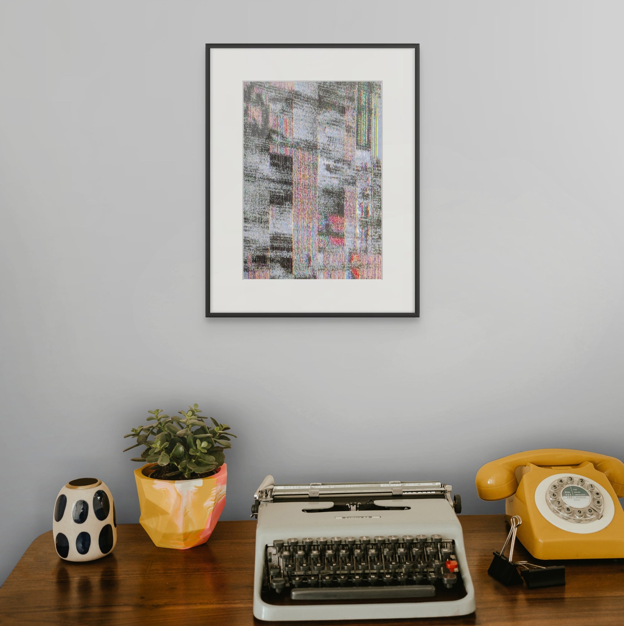 a wooden desk with a typewriter, a yellow rotary phone, and a potted plant. Above the desk, there is a framed abstract painting hanging on the wall.