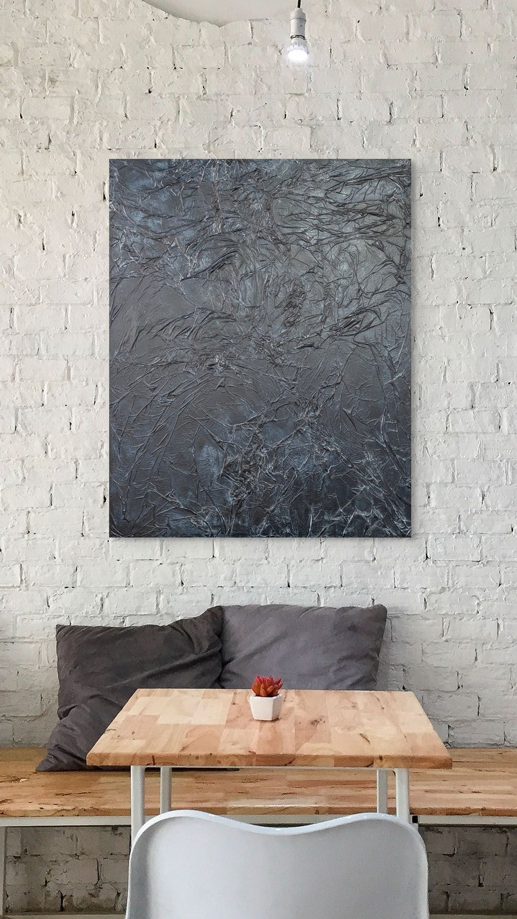 Abstract painting on a white brick wall above a wooden table with a plant and pillows.